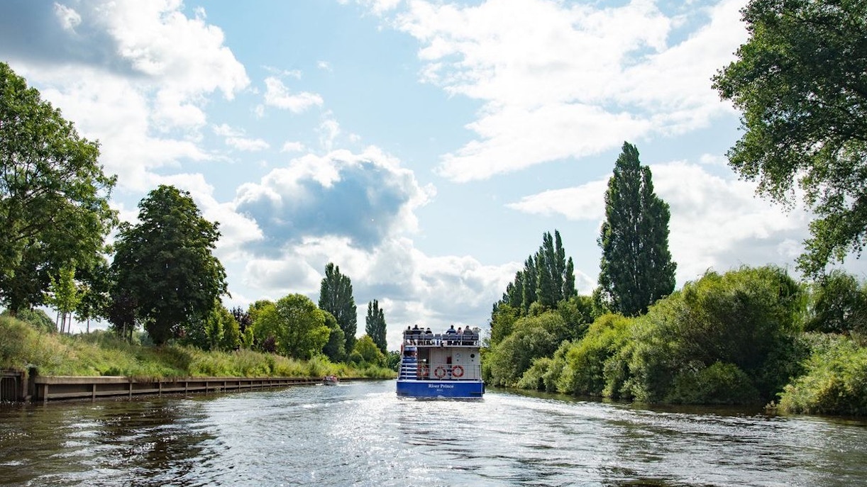 Sightseeing cruise boat on River Ouse in York surrounded by lush greenery.