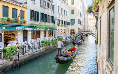 Gondolas navigating a canal in Venice near a restaurant, with a bridge in the background.