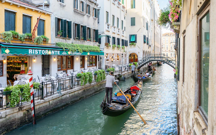 Gondolas navigating a canal in Venice near a restaurant, with a bridge in the background.