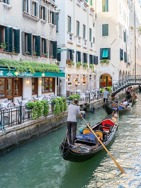 Gondolas navigating a canal in Venice near a restaurant, with a bridge in the background.