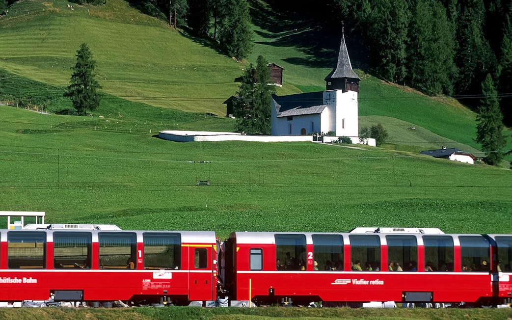 Bernina Express train passing a church in a green landscape between St. Moritz and Tirano.