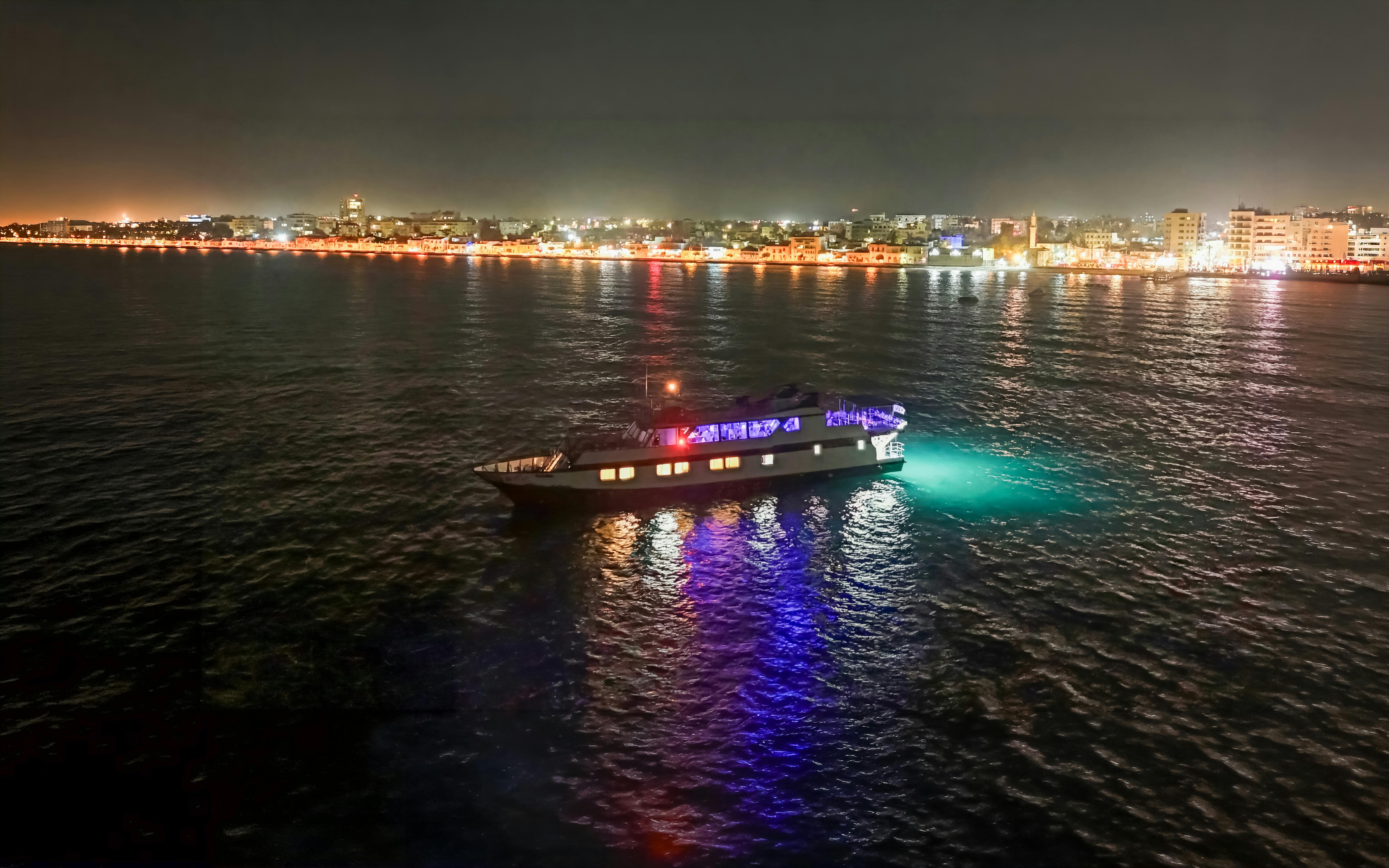 Nighttime disco cruise ship on Larnaca coast with city lights in the background.