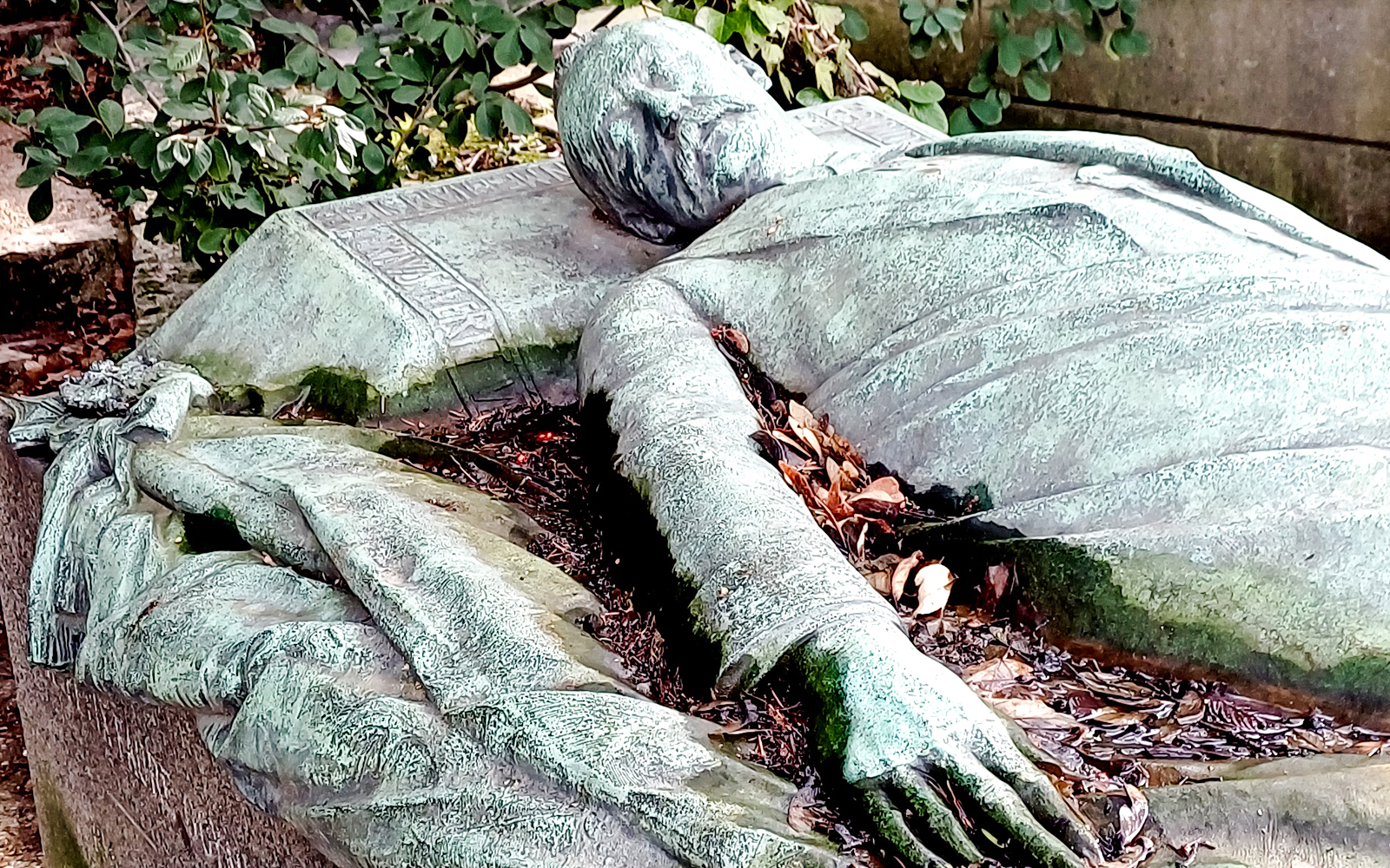 Reclining statue on a tomb in Père Lachaise Cemetery, France, surrounded by foliage.