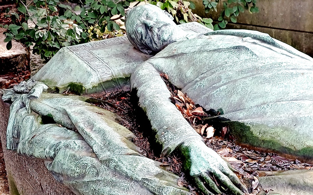 Reclining statue on a tomb in Père Lachaise Cemetery, France, surrounded by foliage.
