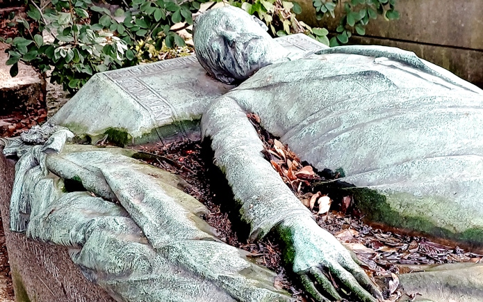 Reclining statue on a tomb in Père Lachaise Cemetery, France, surrounded by foliage.