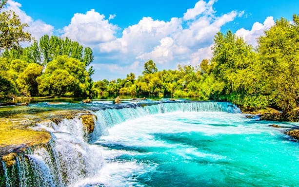 Manavgat River waterfall surrounded by lush greenery under a blue sky.