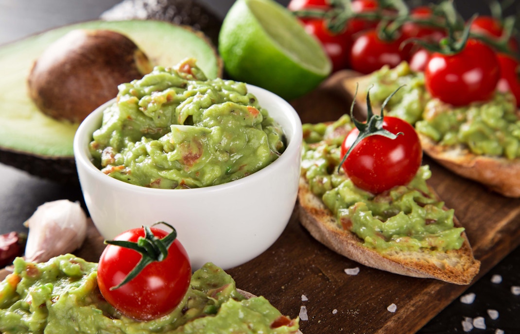 Guacamole in a bowl with cherry tomatoes and avocado toast on a wooden board.