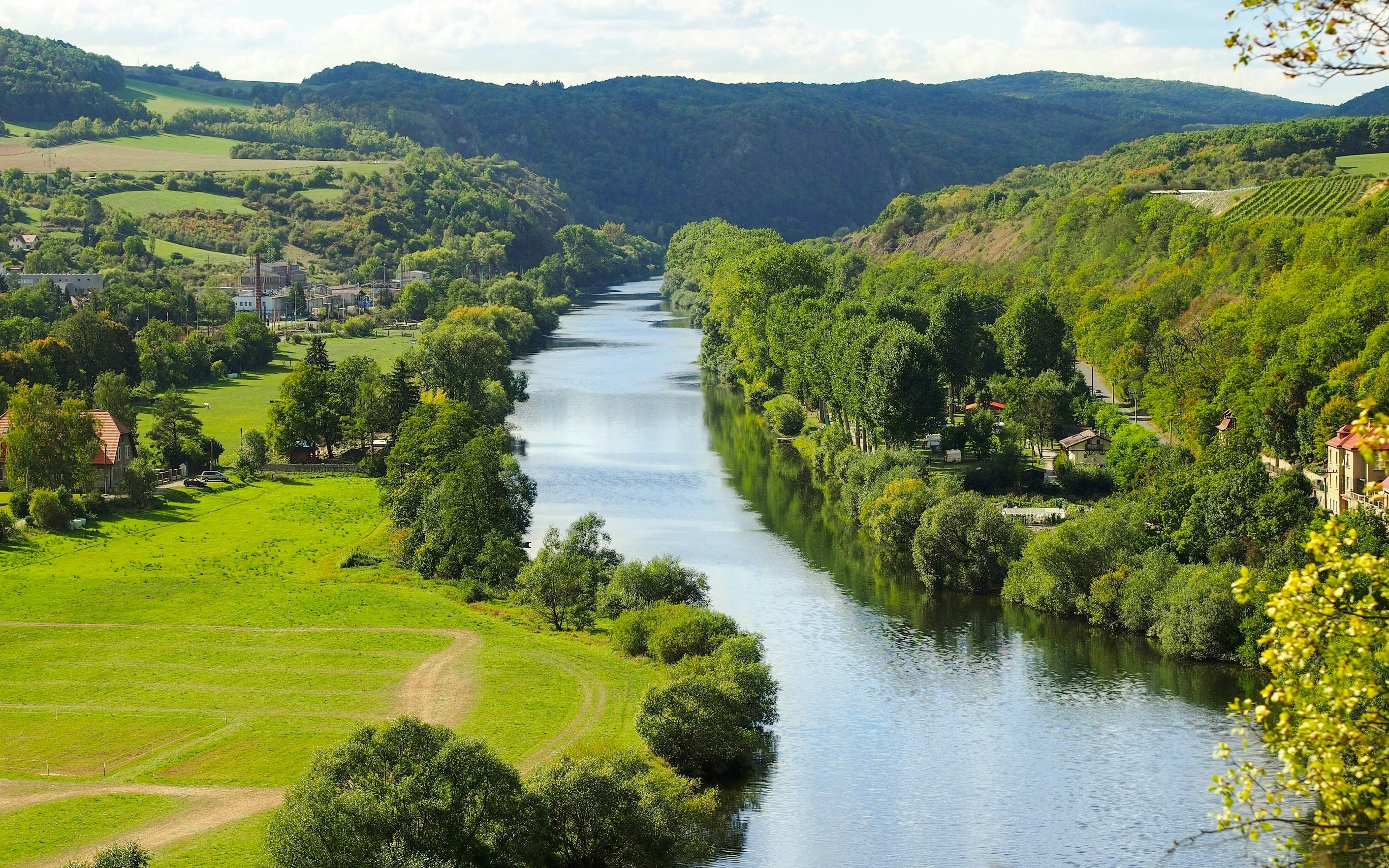 Berounka River flowing through lush landscape near Karlstejn village, surrounded by green hills.