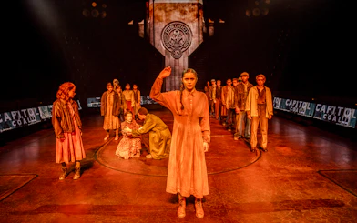 Stage scene from Hunger Games West End show with actors in District 12 setting.