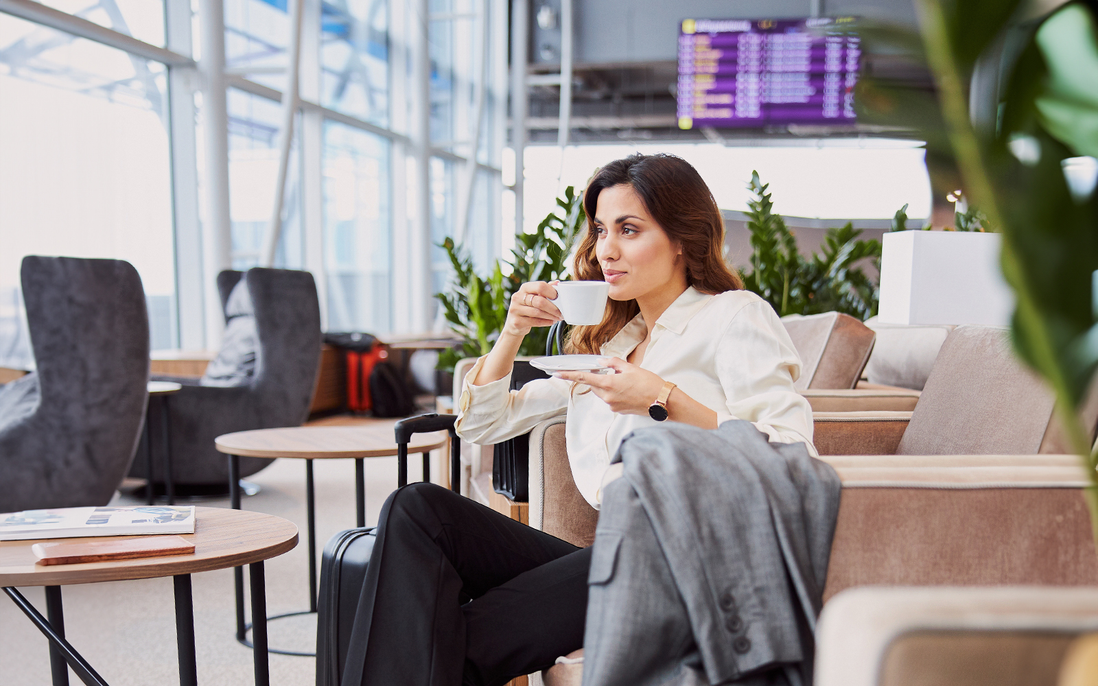 Woman enjoying coffee in airport lounge with flight information display in background.