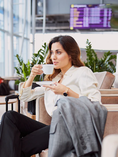 Woman enjoying coffee in airport lounge with flight information display in background.