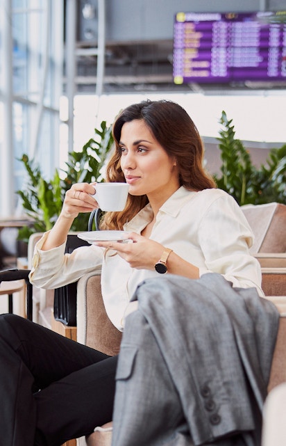 Woman enjoying coffee in airport lounge with flight information display in background.