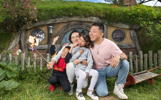 Family taking a selfie in front of a hobbit house at Hobbiton Movie Set, New Zealand.