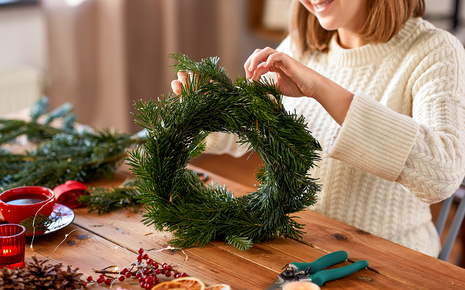 Person crafting a holiday wreath with pine branches on a wooden table.