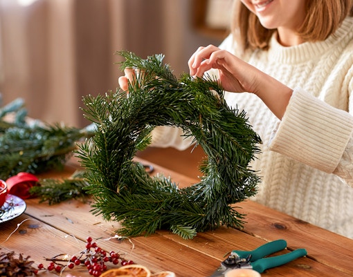Person crafting a holiday wreath with pine branches on a wooden table.
