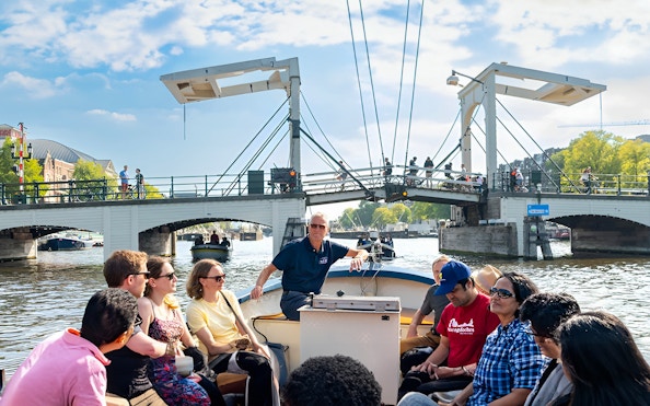 Open boat cruise on Amsterdam canal passing under a drawbridge with passengers enjoying the view.