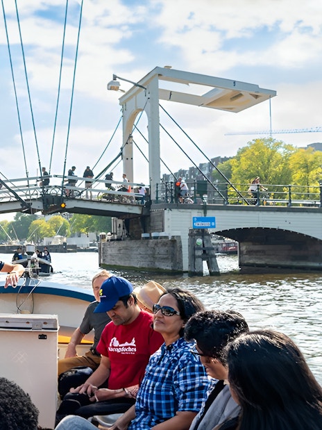 Open boat cruise on Amsterdam canal passing under a drawbridge with passengers enjoying the view.