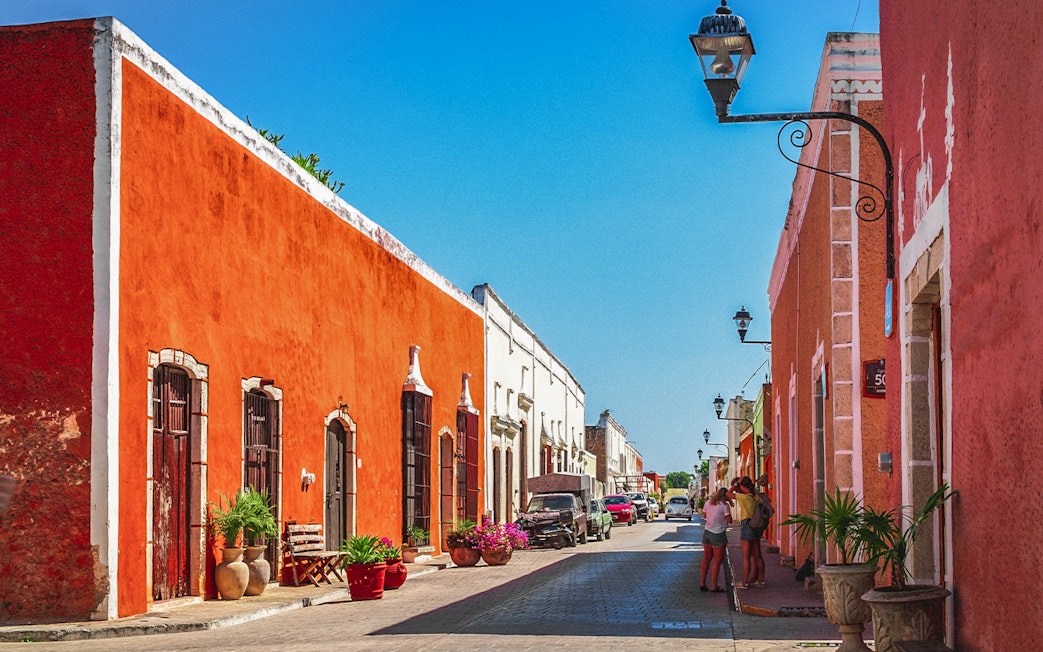 Colorful colonial buildings line a street in Valladolid, Mexico, with people walking and cars parked.