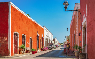 Colorful colonial buildings line a street in Valladolid, Mexico, with people walking and cars parked.