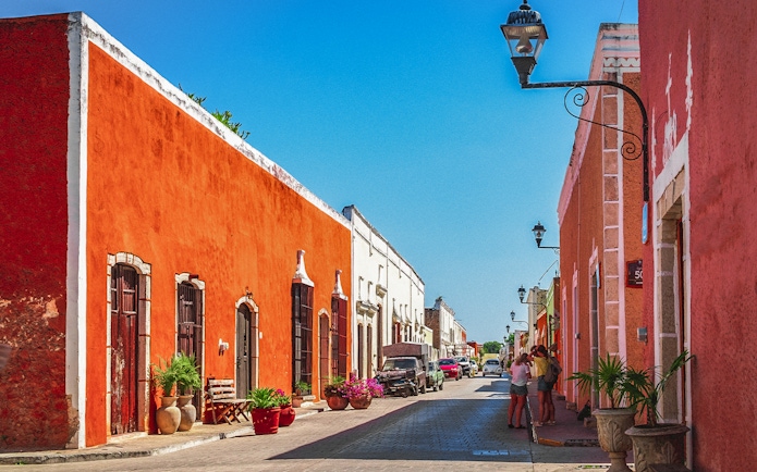 Colorful colonial buildings line a street in Valladolid, Mexico, with people walking and cars parked.