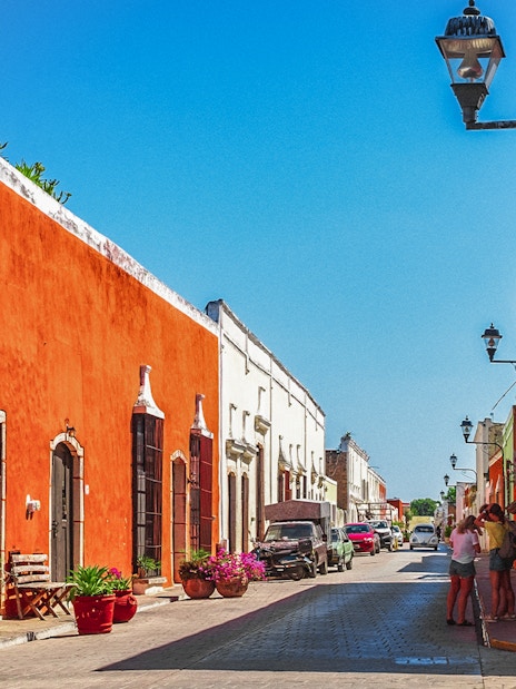 Colorful colonial buildings line a street in Valladolid, Mexico, with people walking and cars parked.