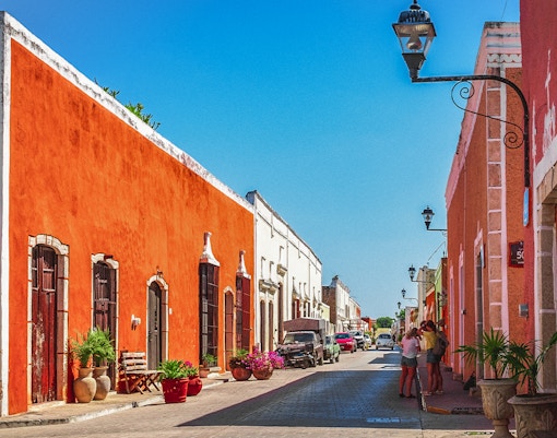 Colorful colonial buildings line a street in Valladolid, Mexico, with people walking and cars parked.