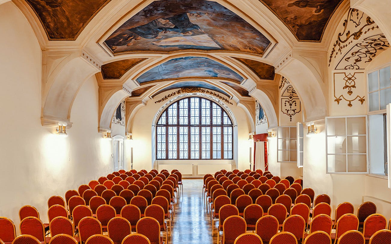 Seating area inside Minorite Monastery of St James, Prague, with ornate ceiling and large window.
