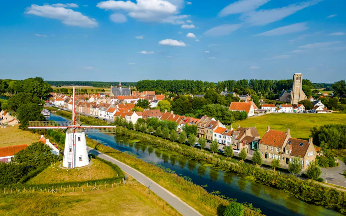 Drone view of Damme, Belgium with windmill, canal, church, and historic buildings under blue sky.