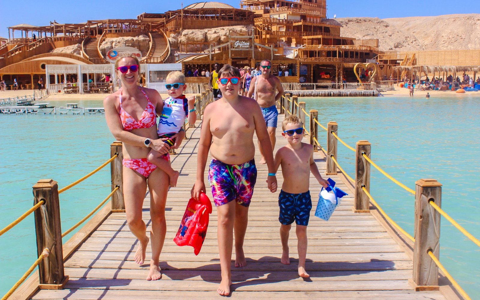 Family walking on a wooden pier at Giftun 3 Islands, with a scenic backdrop of beach and structures.