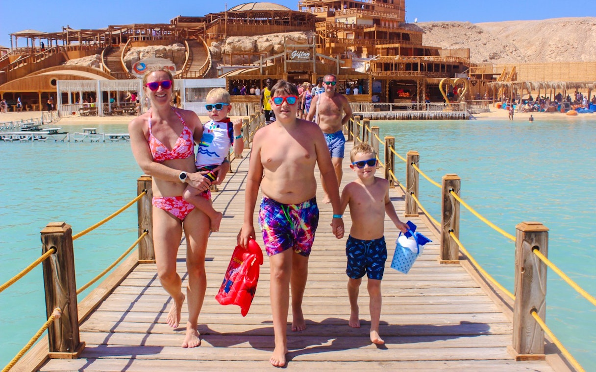 Family walking on a wooden pier at Giftun 3 Islands, with a scenic backdrop of beach and structures.