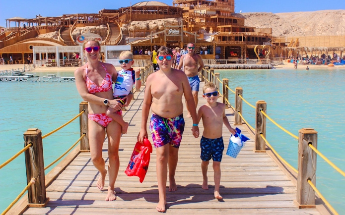 Family walking on a wooden pier at Giftun 3 Islands, with a scenic backdrop of beach and structures.