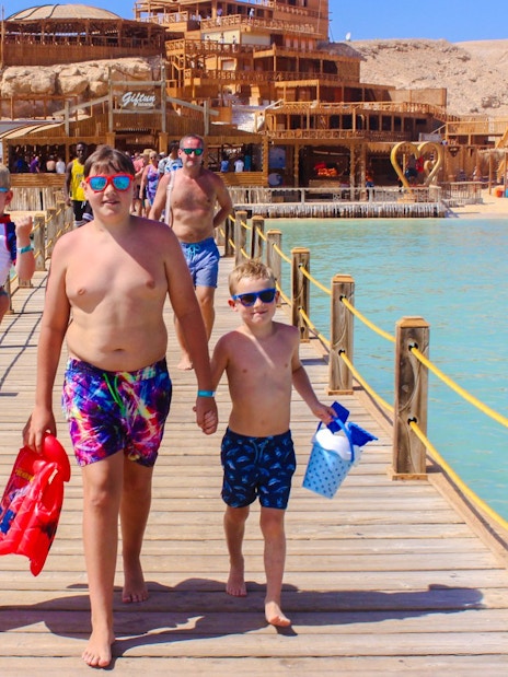 Family walking on a wooden pier at Giftun 3 Islands, with a scenic backdrop of beach and structures.