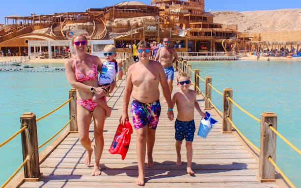 Family walking on a wooden pier at Giftun 3 Islands, with a scenic backdrop of beach and structures.