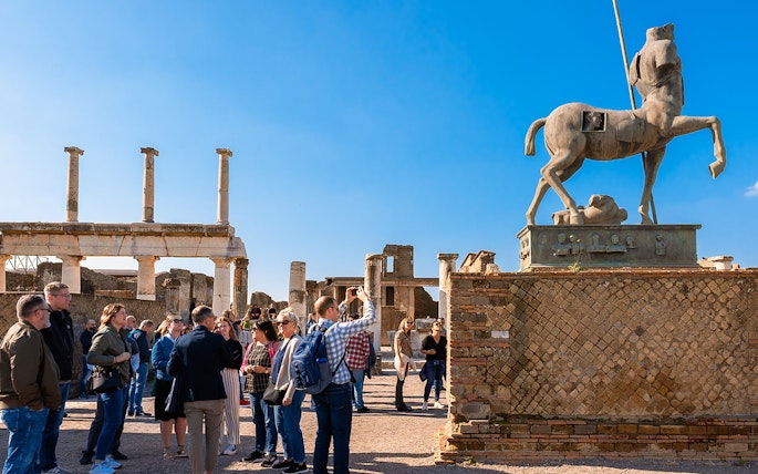 Visitors with a guide exploring the ancient ruins of Pompeii with a centaur statue in view.