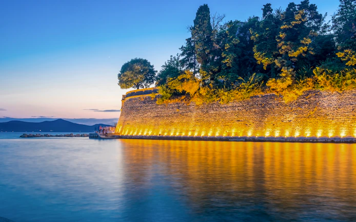 Old city wall in Zadar illuminated at night, reflecting on the water.