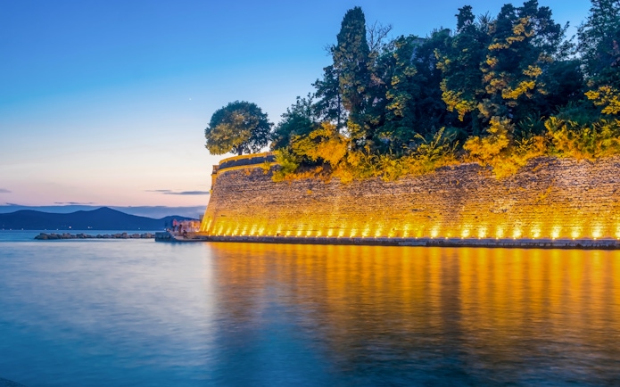 Old city wall in Zadar illuminated at night, reflecting on the water.