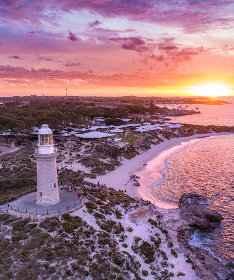Lighthouse on coastal headland at sunset with ocean and pink sky.