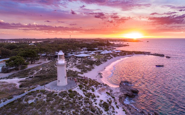 Lighthouse on coastal headland at sunset with ocean and pink sky.