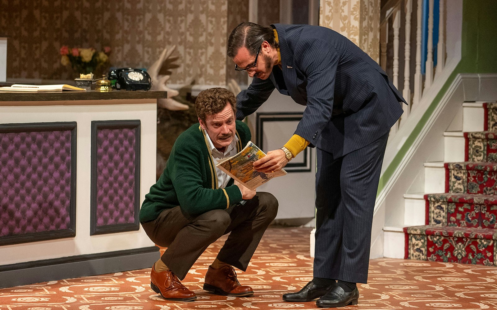 Two actors in a comedic scene at a hotel reception, referencing Fawlty Towers.
