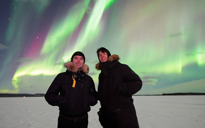 Tourists standing under Northern Lights in snowy landscape.