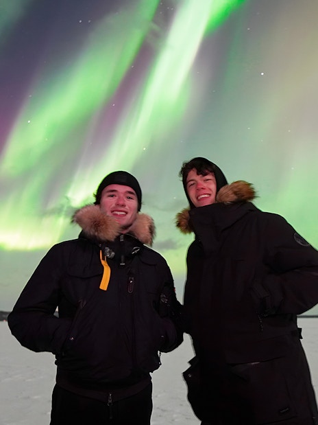 Tourists standing under Northern Lights in snowy landscape.
