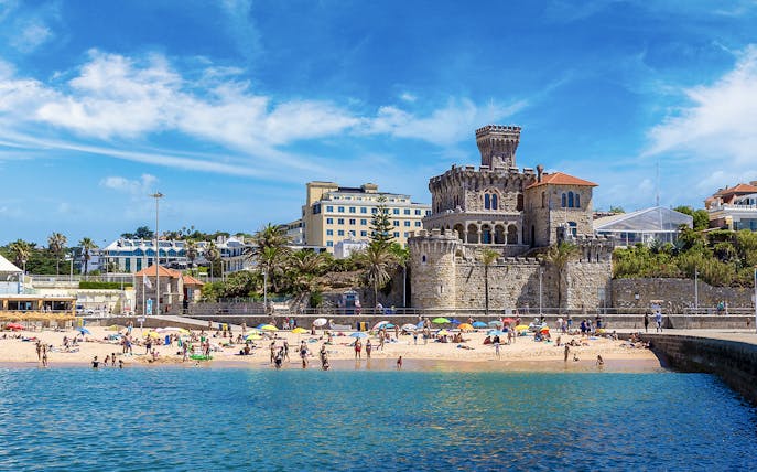 Beachgoers at Cascais with historic castle and buildings in the background, part of Lisbon tour.