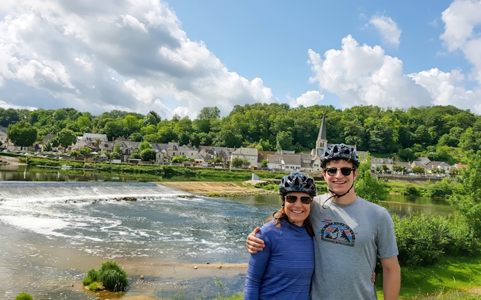 Cyclists by river with village and church in background, full day e-bike tour to Chambord.