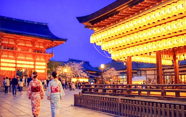 Nighttime view of Yasaka Shrine in Kyoto with illuminated lanterns and visitors in traditional attire.