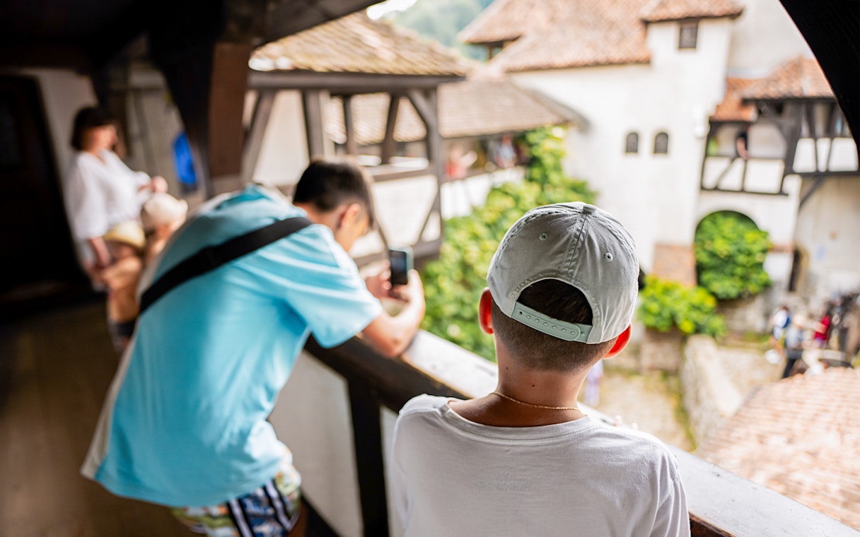 Boy looking into Bran Castle courtyard, capturing the view with a smartphone.