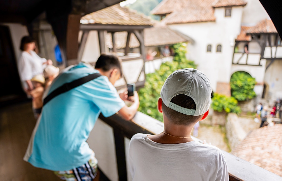 Boy looking into Bran Castle courtyard, capturing the view with a smartphone.