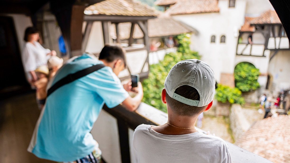 Boy looking into Bran Castle courtyard, capturing the view with a smartphone.
