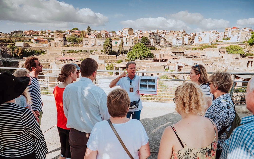 Group listening to a guide at Herculaneum with city ruins in the background.