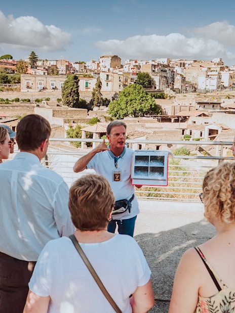 Group listening to a guide at Herculaneum with city ruins in the background.