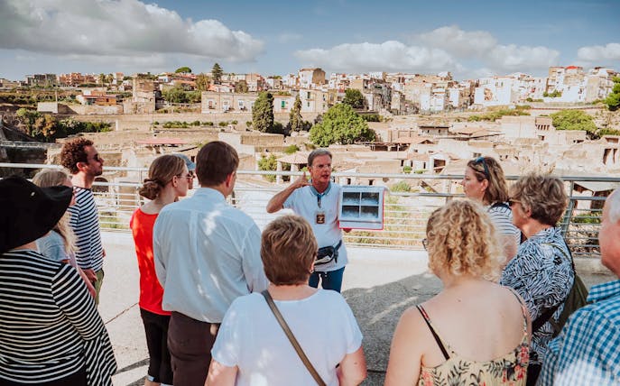 Group listening to a guide at Herculaneum with city ruins in the background.